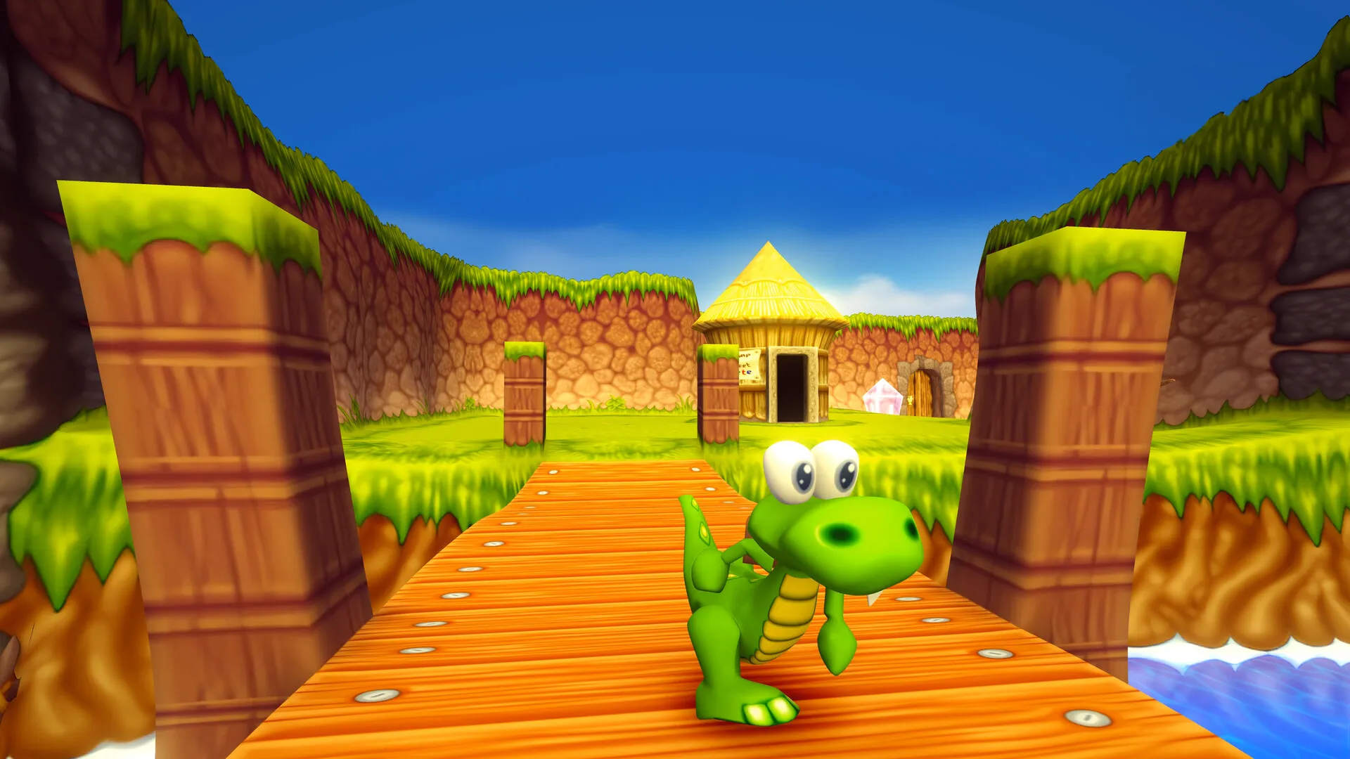 Captura oficial del remaster de Croc 2 Kingdom of the Gobbos: Croc parado en un puente de tablas de madera frente a una cabaña Gobbo con techo de paja y un cristal rosa brillante bajo un cielo azul.
