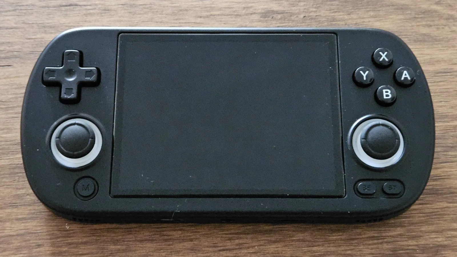 An Anbernic RG40XX handheld, black, with its screen off, lying on a wooden table.