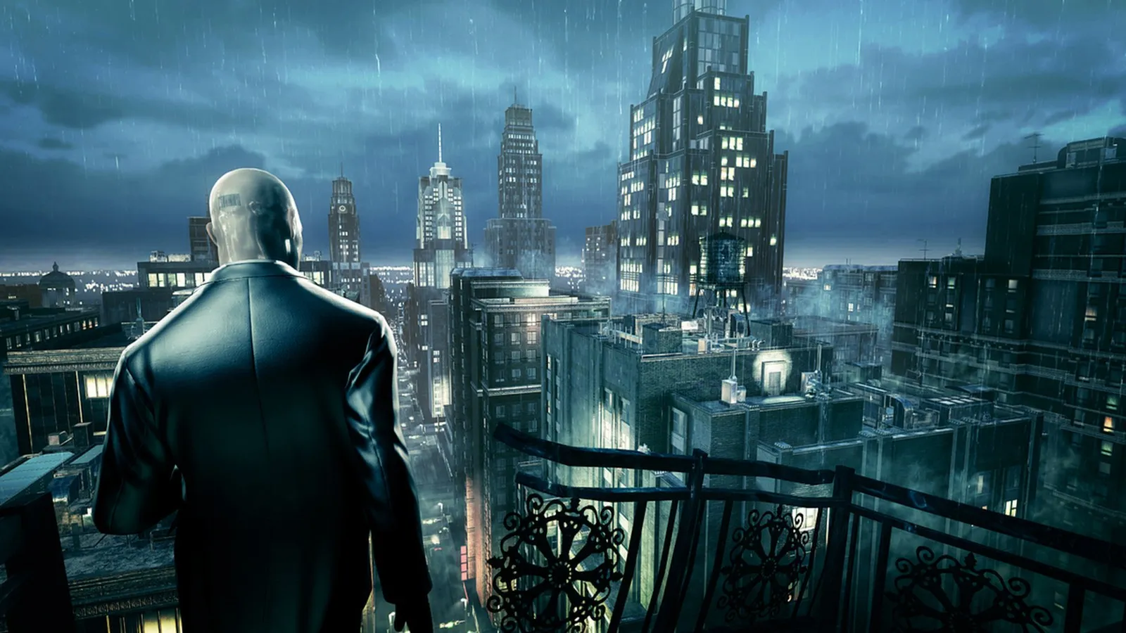 Hitman: Absolution screenshot from the PC release: Agent 47, seen from behind in a dark coat, looks out over a rainy night city skyline from a balcony with ornate ironwork.
