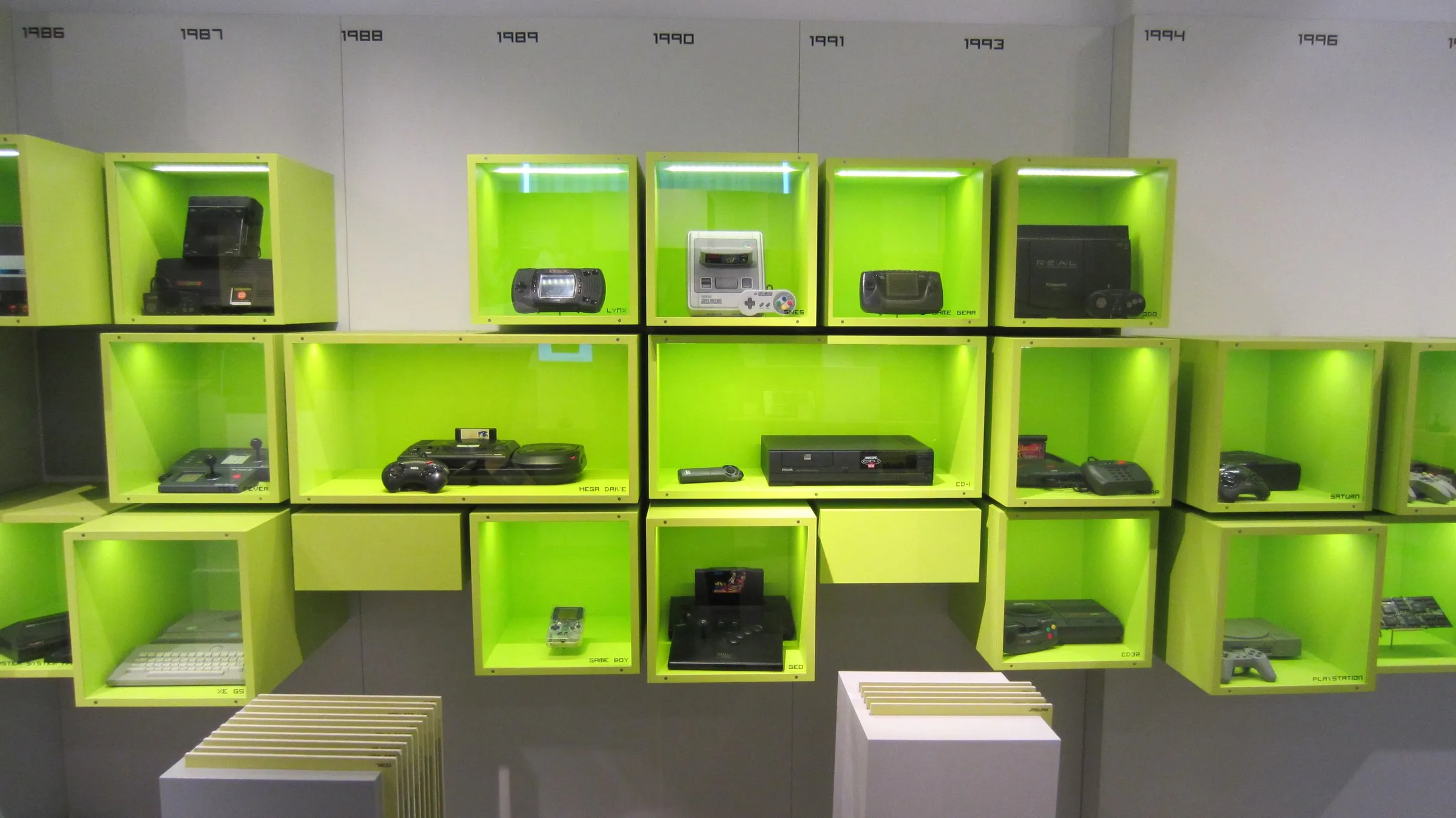 Home consoles from the late 1980s and 1990s displayed one per lit lime-green wall cube, with release years printed along the top of the wall, at the Computerspielemuseum in Berlin.