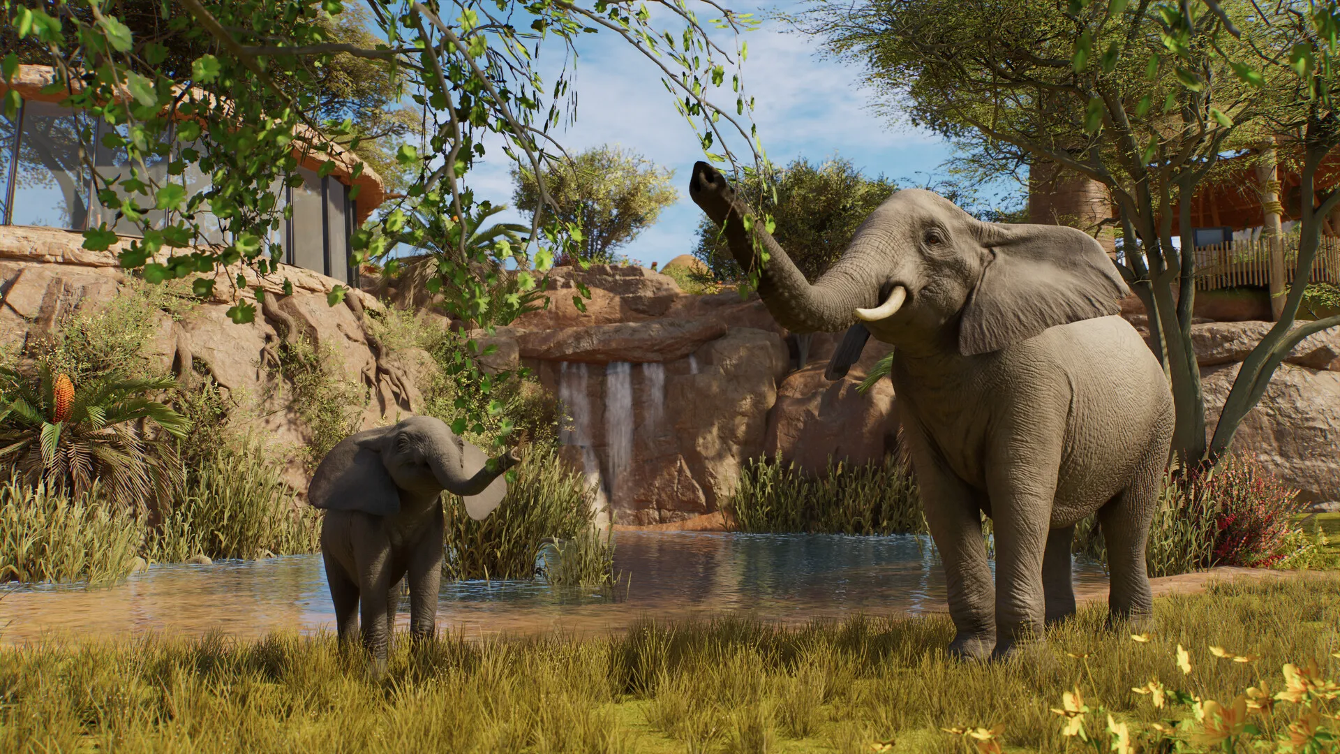 Planet Zoo 2 screenshot: an adult elephant and a calf at a watering hole beneath a rocky waterfall, with a glass visitor building on the left. No interface on screen.
