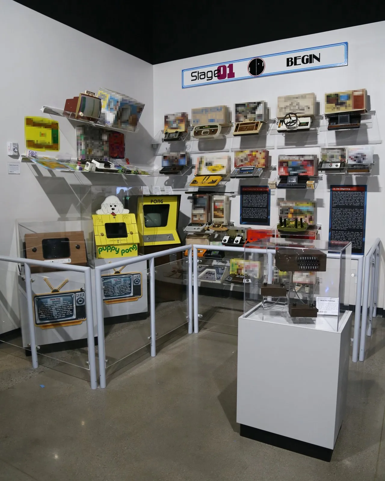 A museum corner of 1970s Pong-era home consoles mounted on a white wall and in glass cases, under a sign reading “Stage 01 Begin.”